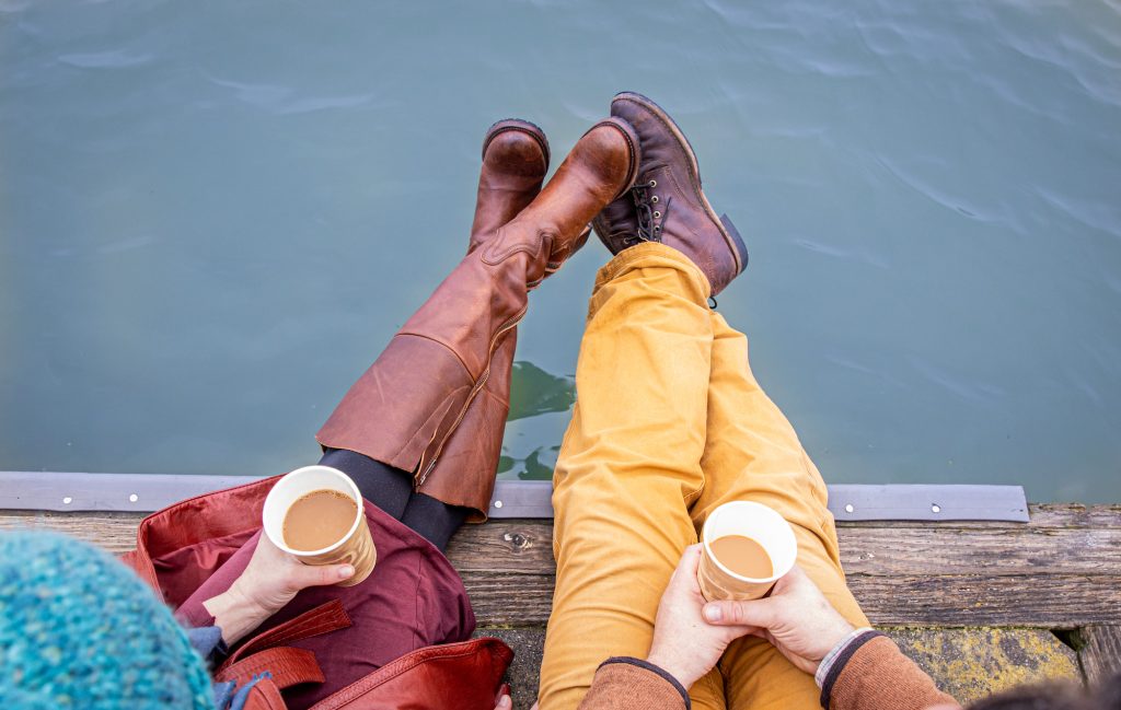 Couple holding coffee sitting on a dock with their feet over the water